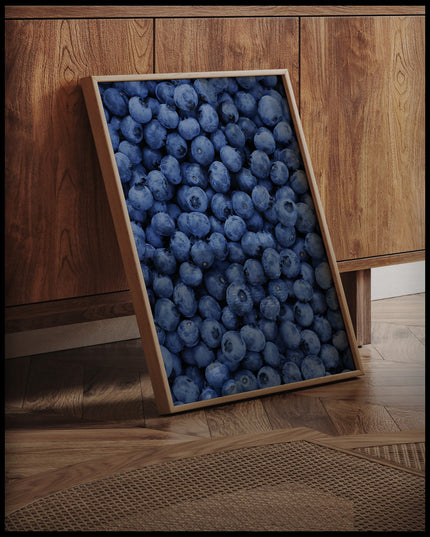 Ein gerahmtes Poster von einer Fülle von frischen Blaubeeren, die das gesamte Bild bedecken, an einer Holzkommode angelehnt und stehend auf dem Holzboden