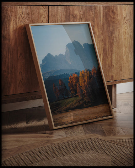 Ein gerahmtes Poster von herbstlich gefärbten Bäumen vor einer Berglandschaft in den Dolomiten, an einer Holzkommode angelehnt und stehend auf dem Holzboden