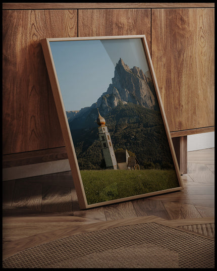 Ein gerahmtes Poster von einer Kirche am Abhang eines Berges in den Dolomiten, an einer Holzkommode angelehnt und stehend auf dem Holzboden