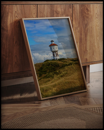 Ein gerahmtes Poster von einem weißen Wasserturm auf der Insel Langeoog, an einer Holzkommode angelehnt und stehend auf dem Holzboden