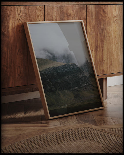 Ein gerahmtes Poster von einem steilen Abhang eines Berges, der von Gras bedeckt und von Nebel umschlungen ist, an einer Holzkommode angelehnt und stehend auf dem Holzboden