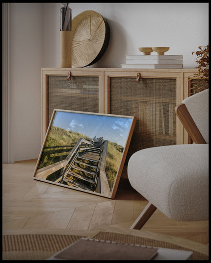 Ein gerahmtes Poster von einer Holztreppe die über Dünen am Strand führt, an einer Holzkommode angelehnt und stehend auf dem Holzboden