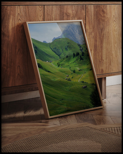 Ein gerahmtes Poster von einer grünen Berglandschaft mit einzelnen Bäumen und Holzhütten, an einer Holzkommode angelehnt und stehend auf dem Holzboden