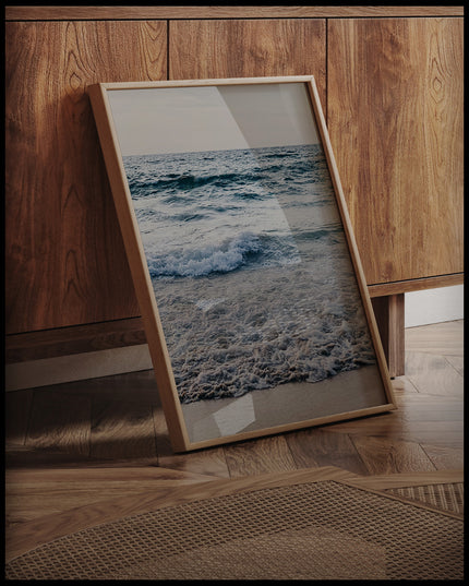 Ein gerahmtes Poster von schaumigen Wellen am Strand, an einer Holzkommode angelehnt und stehend auf dem Holzboden