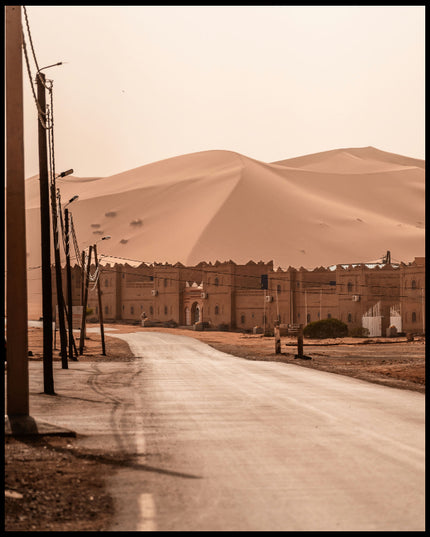 Ein Poster von einer Straße, die zu einer Siedlung am Rande einer weitläufigen Wüstenlandschaft mit hohen Sanddünen führt.