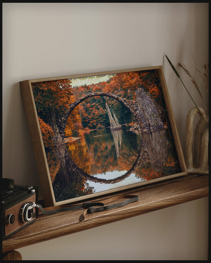 Ein gerahmtes Poster von der Seitenansicht einer freispannenden Bogenbrücke in einem herbstlichen Wald und über einem ruhigen See, auf einem Holzregal an die Wand angelehnt