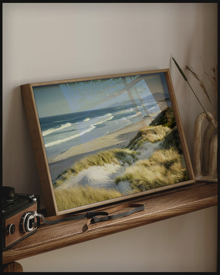 Ein gerahmtes Poster vom Meerblick über Dünen und Strand, auf einem Holzregal an die Wand angelehnt