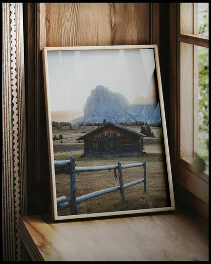 Ein gerahmtes Poster von einer rustikalen Hütte in einer herbstlichen Berglandschaft, vor einem Fenster an die Wand angelehnt