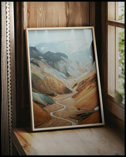 Ein gerahmtes Poster von einem Tal mit einem Fluss und orangenen Bergen, vor einem Fenster an die Wand angelehnt