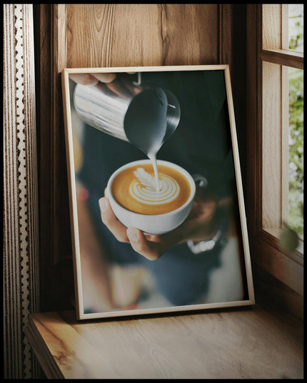 Ein gerahmtes Poster von einem Barista, der kunstvoll Milch in eine Tasse Kaffee gießt und ein Muster kreiert, vor einem Fenster an die Wand angelehnt