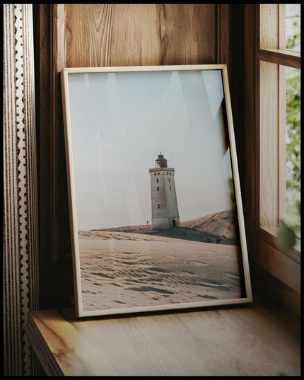 Ein gerahmtes Poster von einem einsamen Leuchtturm in einer sandigen Dünenlandschaft unter einem klaren Himmel, vor einem Fenster an die Wand angelehnt