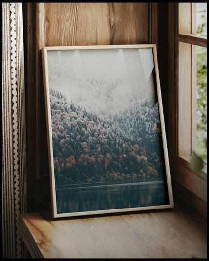 Ein gerahmtes Poster von einem nebeligen Nadelwald, der stellenweise von Schnee bedeckt ist und an einen See grenzt, vor einem Fenster an die Wand angelehnt