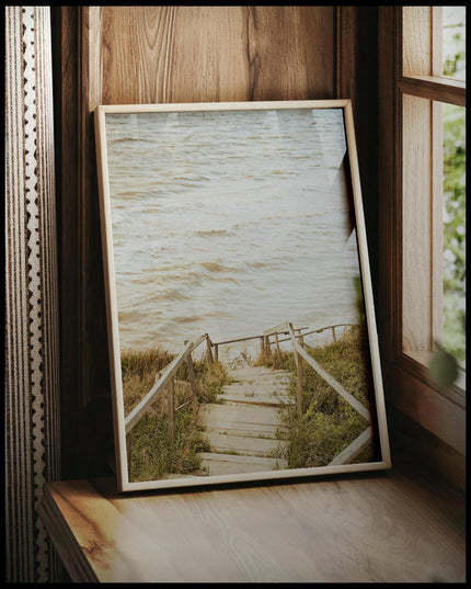 Ein gerahmtes Poster von einer Holztreppe die durch Dünen runter zum Strand führt, vor einem Fenster an die Wand angelehnt