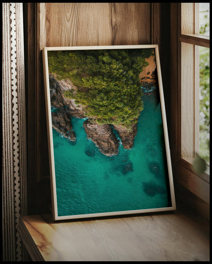 Ein gerahmtes Poster von einer steilen, bewachsenen Klippe und blauem Meer, vor einem Fenster an die Wand angelehnt